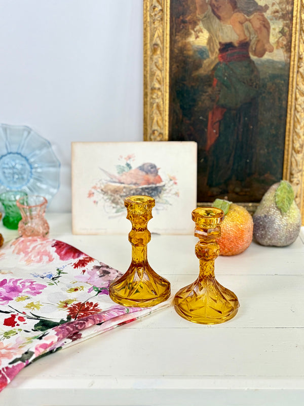 Vintage amber pressed glass candlestick pair on white cupboard beside floral linen cloth with vintage art and objects in background