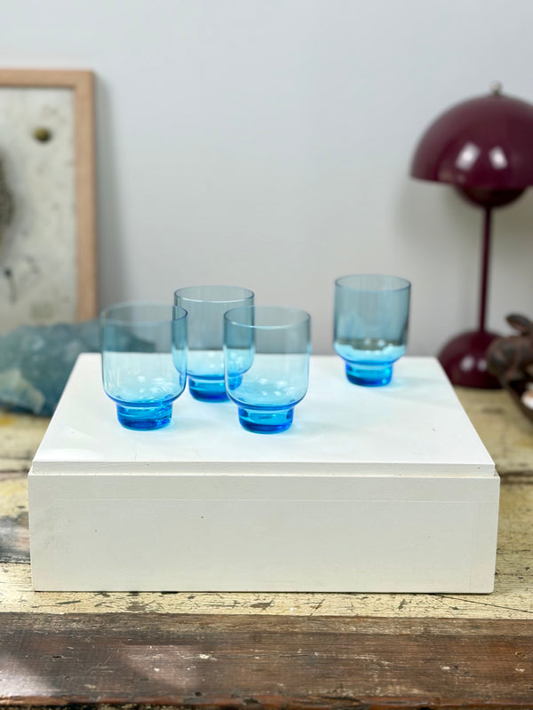 Closer view of three Crown Crystal Mondial Blue ice blue tumblers on white plinth with fourth spare tumbler to the side, timber table with decorative objects and grey wall background
