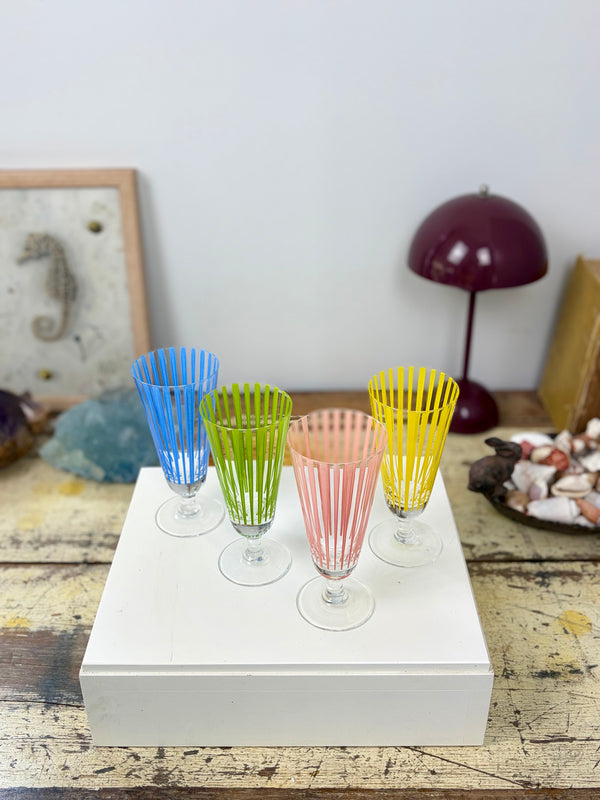 High angle view of four vintage striped stemmed glasses in blue, green, pink and yellow on white plinth on timber table with decorative objects