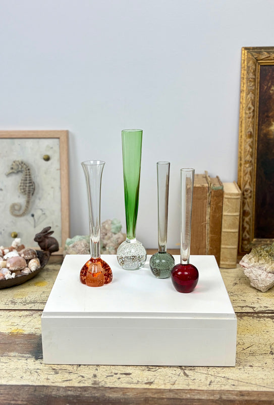Five decorative glass vases in assorted colors and shapes displayed on a white rectangular box on a rustic wooden table, with framed art and books in the background.