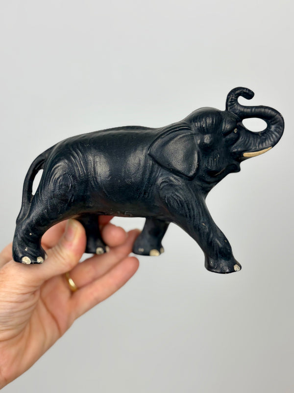 SylvaC Model 815 matte black elephant figurine held from underneath in an open hand, full right profile view against plain grey background — trunk raised, single white tusk visible, incised skin and wrinkle detail across body, white-tipped toes on all four feet, scale reference