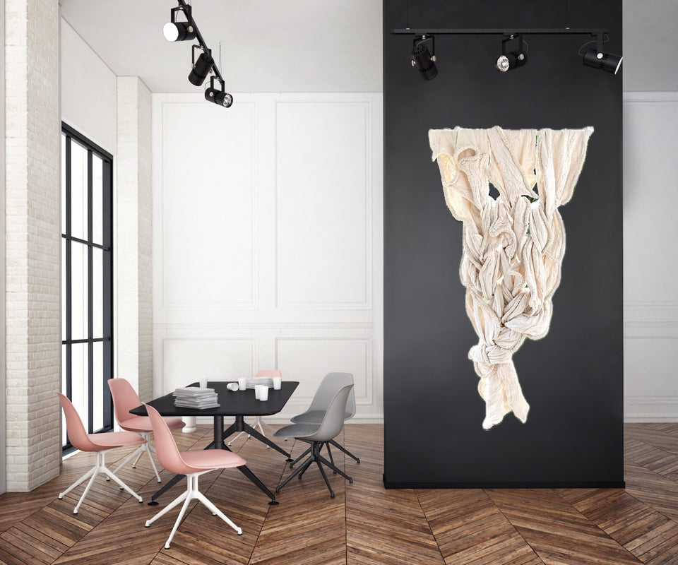A modern interior room with a black wall panel displaying a large, sculptural, draped cream-colored textile art piece. The room features a wooden herringbone floor, a dining table, and pink and gray chairs.