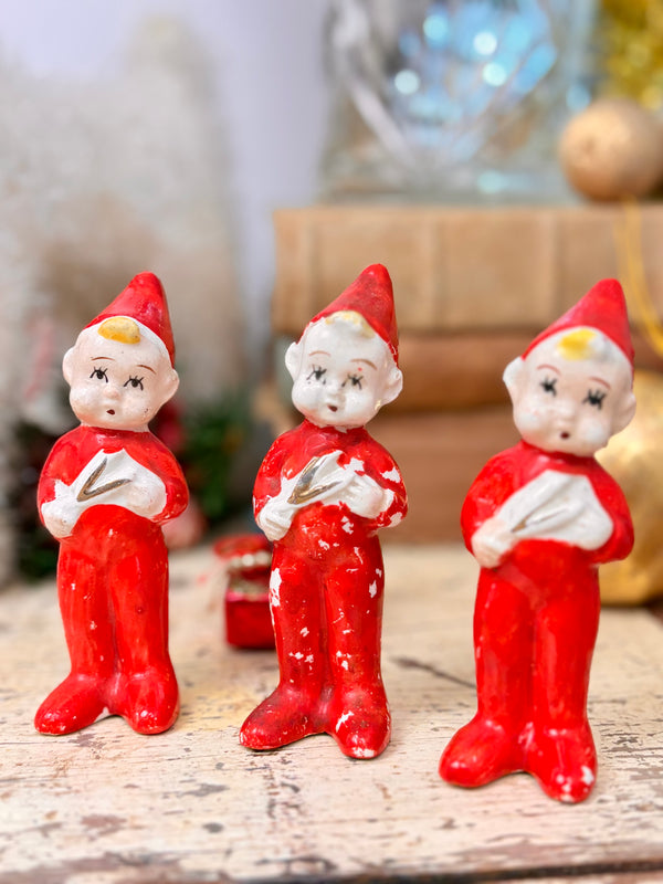 Three vintage Christmas caroller figurines holding white songbooks, with red pointed hats, painted faces and visible wear to their red suits.