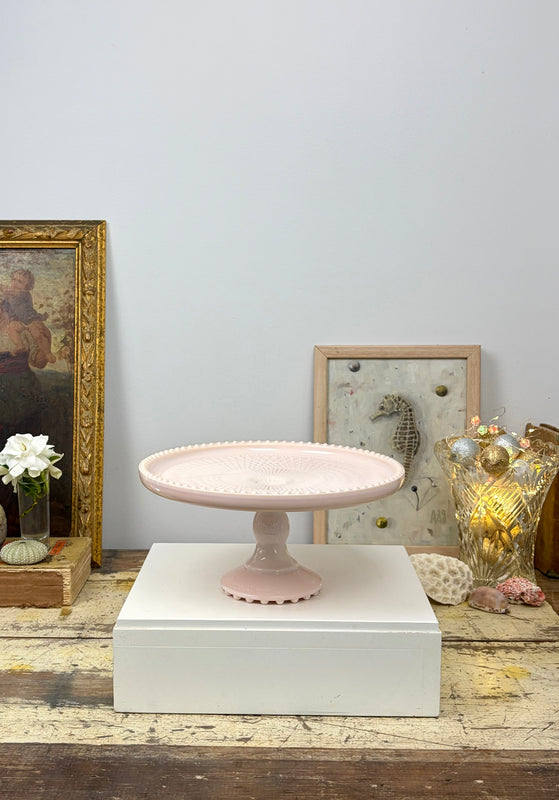 Jeannette Shell Pink milk glass cake stand on a white plinth, styled with framed artwork, shells and vintage glass.