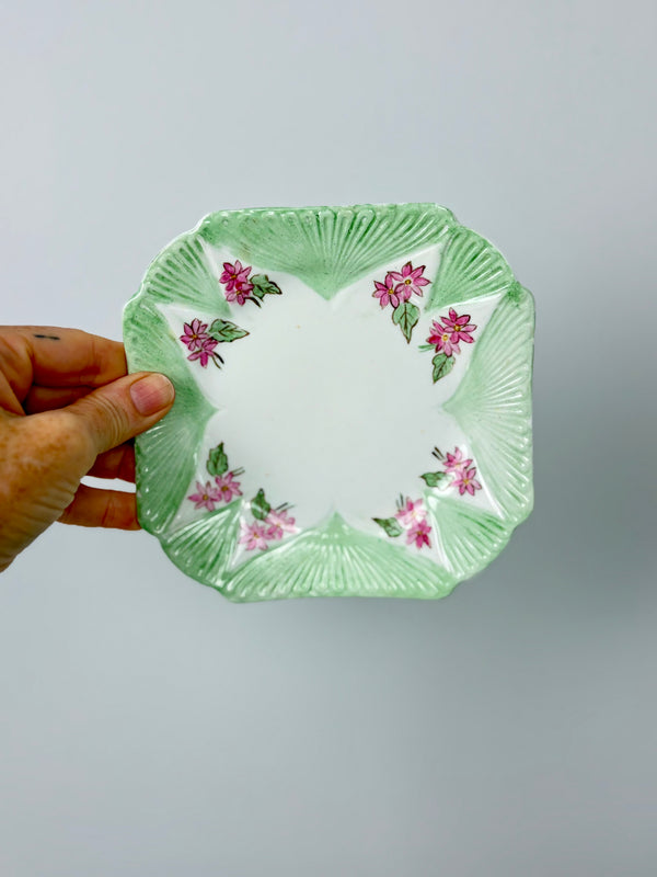 Hand holding Shelley Dainty Square seafoam side plate vertically against grey wall showing hand-painted pink starburst floral and foliage detail
